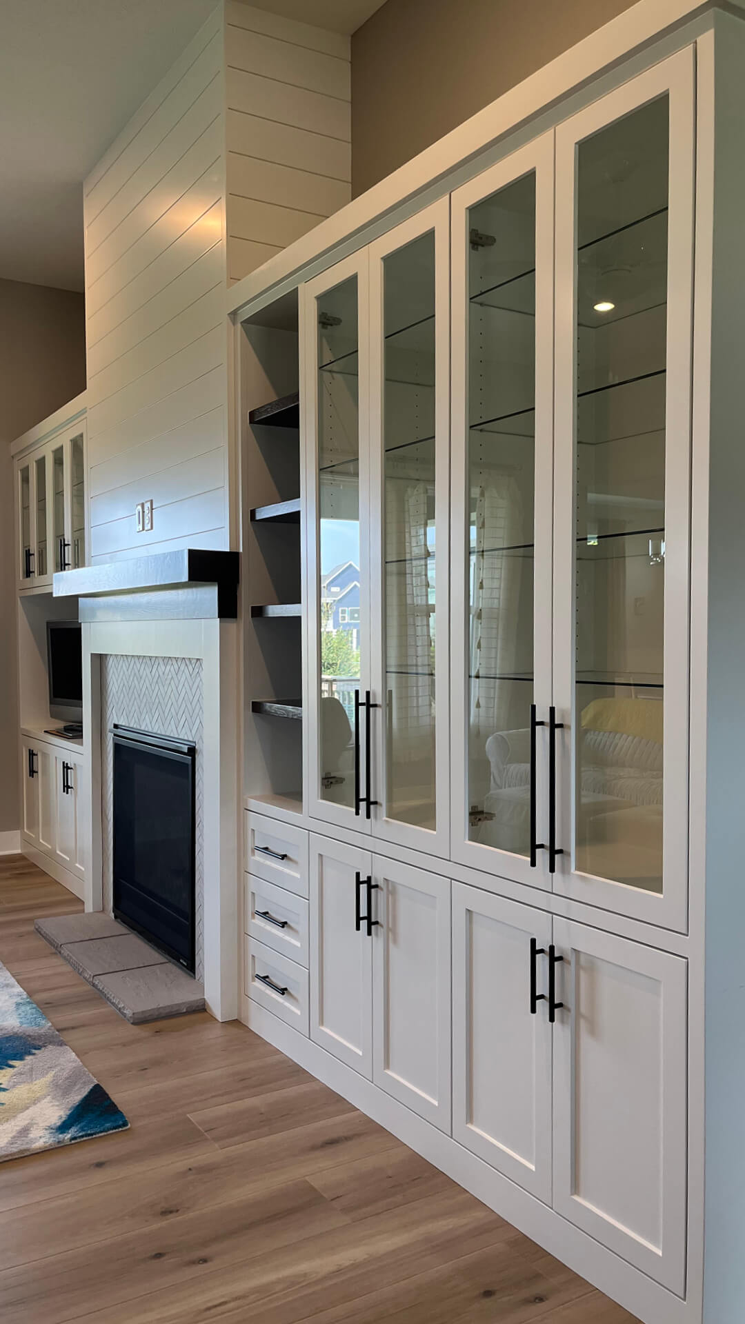 Custom white cabinets with glass doors and black handles in a room with wooden flooring.